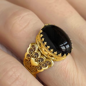 Puede incluir: Un anillo de oro con una piedra ovalada negra engastada en un diseño de filigrana. El anillo presenta un diseño en forma de corazón en la banda.