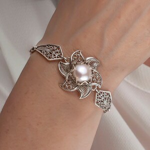 May include: A silver bracelet with a delicate filigree design features a large, white pearl in the center. The bracelet is on a woman's wrist.