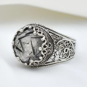 以下が含まれることがあります： 黒いインクルージョンが入った、楕円形の透明な宝石が付いたシルバーリング。リングは、石の周りとバンドに、装飾的なフィリグリーデザインが施されています。リングは白い背景に配置されています。