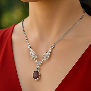 Puede incluir: Un collar de plata con un colgante ovalado rojo oscuro. El collar tiene una cadena delicada y un diseño geométrico adornado con pequeñas piedras transparentes. La mujer lleva un vestido rojo.