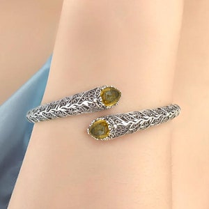 Op de afbeelding: Een zilveren armband met een delicaat, bladerig ontwerp. De armband heeft twee peervormige edelstenen, één aan elk uiteinde, met een geelgroene tint.