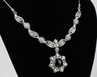 May include: A silver filigree necklace with a black gemstone pendant. The necklace is displayed on a black velvet stand.