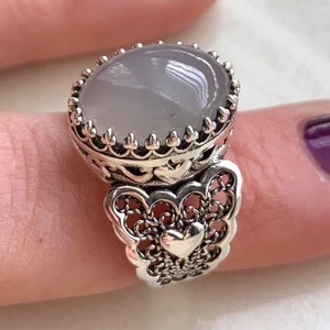 May include: A silver ring with a large oval-shaped grey stone set in a filigree design. The ring has a heart-shaped design on the band.