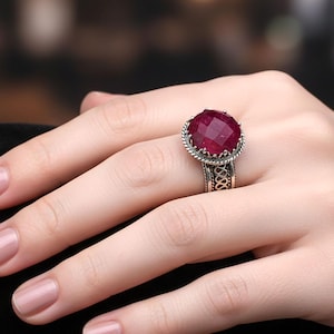 May include: A silver ring with a large, faceted, oval-shaped ruby gemstone. The ring has an ornate design around the gemstone and on the band. The ruby is a deep red color, and the ring is worn on a finger.