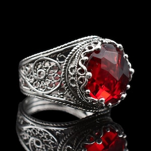 Puede incluir: Un anillo de plata con un intrincado diseño de filigrana y una gran gema roja facetada. El anillo se muestra sobre un fondo negro, con su reflejo visible debajo. La gema roja es el punto focal, captando la luz y añadiendo color.