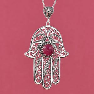 May include: Silver pendant necklace with a filigree Hamsa hand design. The pendant features a central, round, red gemstone. The necklace chain is delicate and silver. The background is a solid, muted red.