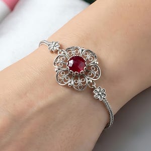 May include: A silver bracelet featuring a filigree flower design with a large, round, red gemstone center. The bracelet is delicate and detailed, with small flower accents along the chain. The jewelry is displayed on a person's wrist.