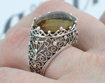 May include: A silver ring with an oval, amber-colored gemstone. The ring features an intricate filigree design around the gemstone and band. The ring is worn on a finger, showcasing the detailed craftsmanship and the gemstone's color.
