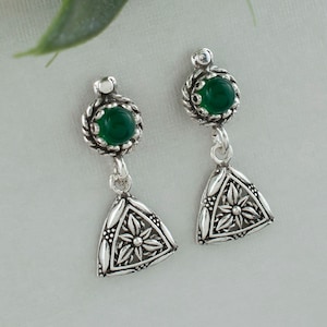 May include: A pair of silver earrings with green gemstone accents. Each earring features a round green stone set in a silver frame, followed by a dangling triangular charm with a floral design.