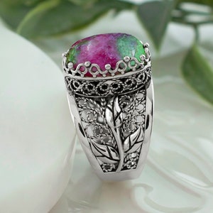 May include: A silver ring featuring an oval green and pink gemstone set in a filigree design. The ring has a floral motif on the band.