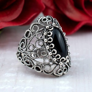 May include: A silver filigree ring with a black oval gemstone. The ring is set against a background of red roses.