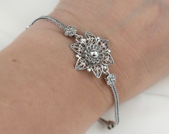 May include: A silver bracelet with a delicate filigree flower charm. The charm has a small bead in the center and is connected to the chain by two smaller flower charms.