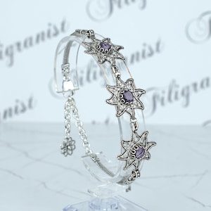 May include: A silver bracelet with a filigree design and purple gemstones. The bracelet is on a clear display stand.