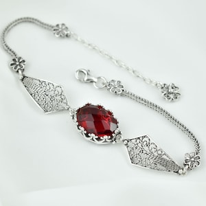 May include: A silver bracelet with a large, oval, faceted red gemstone as the centerpiece. The bracelet features intricate filigree diamond-shaped links and delicate floral accents. The chain is adjustable.