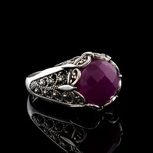 May include: A silver ring with a large, faceted, deep purple gemstone. The ring has an ornate, filigree design around the gemstone and on the band. The ring is set against a black background, with a reflection visible below.