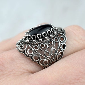 May include: A silver filigree ring with a black oval gemstone. The ring is intricate and delicate, with a detailed design.