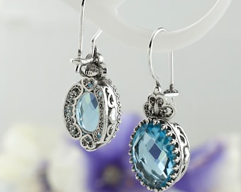 May include: A pair of silver earrings with blue gemstone accents. The earrings feature a delicate filigree design and a round, faceted gemstone.