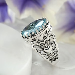 May include: A silver ring with a large, oval-shaped blue gemstone set in a filigree design. The ring features intricate details and a heart-shaped design on the band.