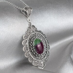 May include: A silver filigree pendant necklace with a green and purple gemstone. The pendant is oval shaped and has a delicate, intricate design.