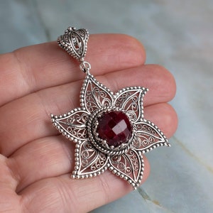 May include: A silver filigree pendant with a red gemstone in the center. The pendant is shaped like a star and has a delicate, intricate design.