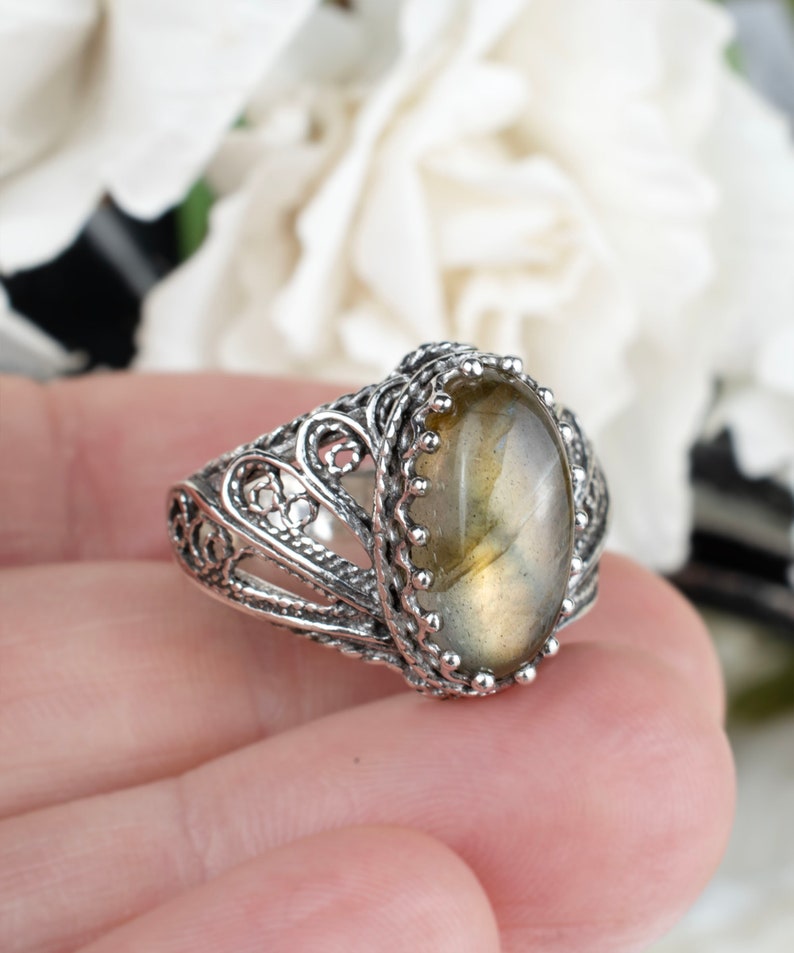 May include: A silver ring with an oval, light-colored gemstone. The ring features an ornate, filigree design around the stone and band. The gemstone is set in a detailed bezel. The ring is held in a person's hand, with a blurred floral background.