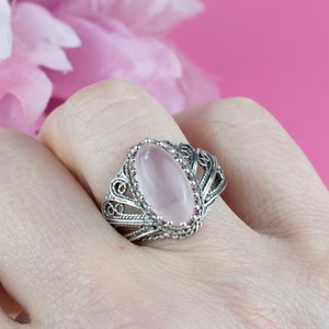 May include: A silver filigree ring with an oval, light pink gemstone. The ring's intricate design features delicate swirls and patterns around the stone, set against a soft pink background. A close-up shot highlights the ring's craftsmanship.