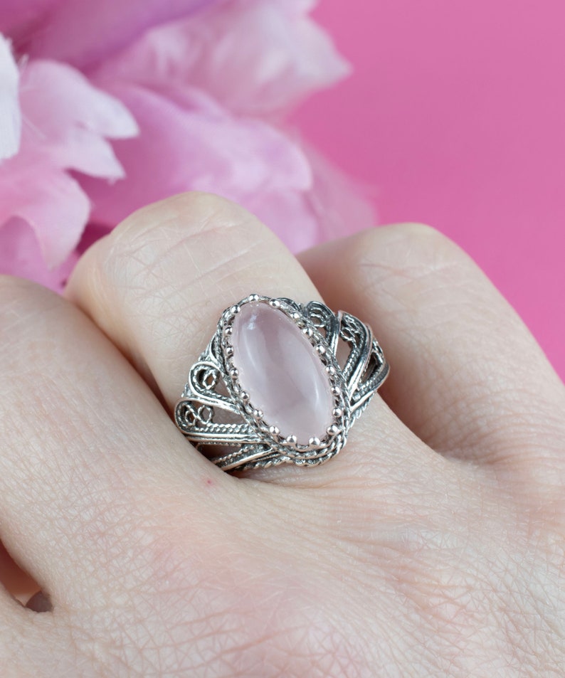 May include: A silver filigree ring with an oval, light pink gemstone. The ring's intricate design features delicate swirls and patterns around the stone, set against a soft pink background. A close-up shot highlights the ring's craftsmanship.