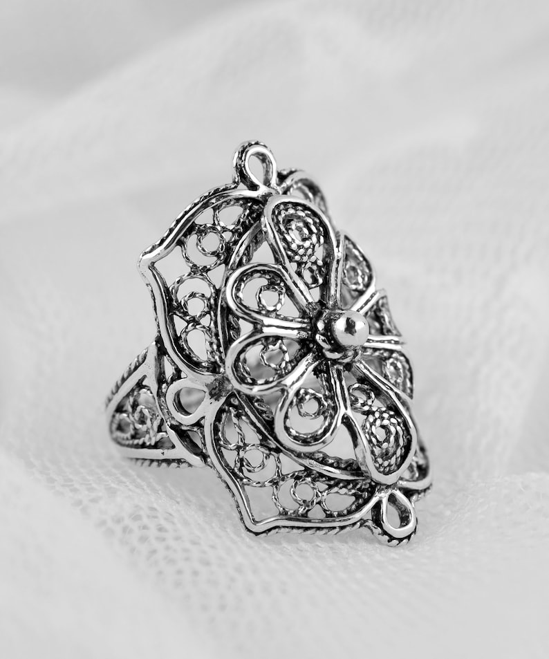 May include: A silver filigree ring with a floral design. The ring is delicate and intricate, with a small bead at the center of the flower.