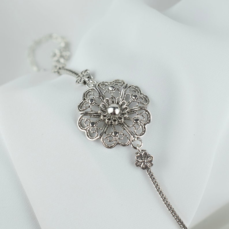 May include: A silver bracelet with a delicate filigree flower charm. The charm has a round center and intricate details.