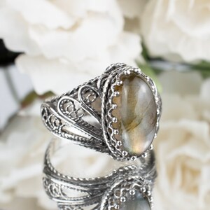 May include: A silver ring with an oval, iridescent gemstone. The ring features an ornate, filigree design around the band and the setting of the stone. The gemstone has a light green and yellow color. The ring is displayed on a reflective surface.
