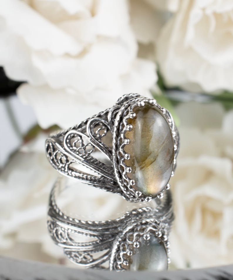 May include: A silver ring with an oval, iridescent gemstone. The ring features an ornate, filigree design around the band and the setting of the stone. The gemstone has a light green and yellow color. The ring is displayed on a reflective surface.