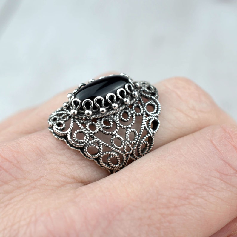 May include: A silver filigree ring with a black oval gemstone. The ring is intricate and delicate, with a detailed design.