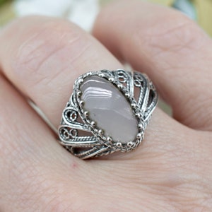 May include: A silver filigree ring featuring a light pink oval gemstone. The ring has an ornate design with detailed metalwork surrounding the stone. The ring is a piece of jewelry.