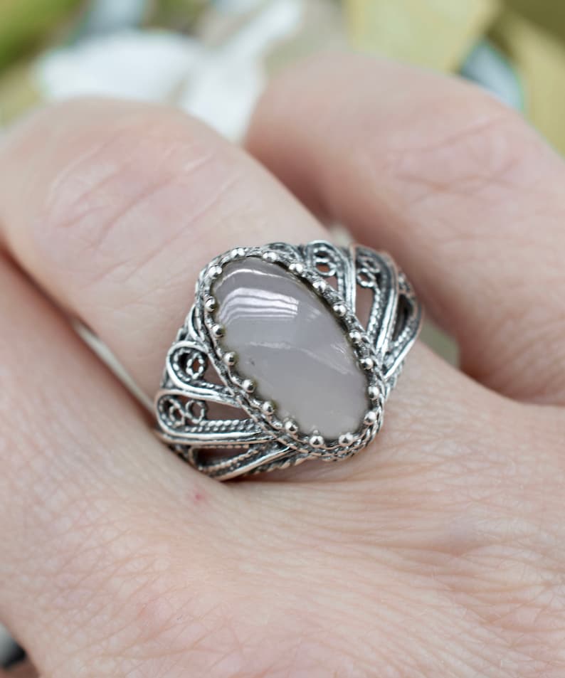 May include: A silver filigree ring featuring a light pink oval gemstone. The ring has an ornate design with detailed metalwork surrounding the stone. The ring is a piece of jewelry.
