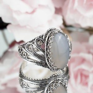 May include: A silver ring with an oval, gray gemstone. The ring has an ornate, filigree design around the band and the gemstone setting. The ring is reflected in a mirrored surface, with soft pink flowers in the background.