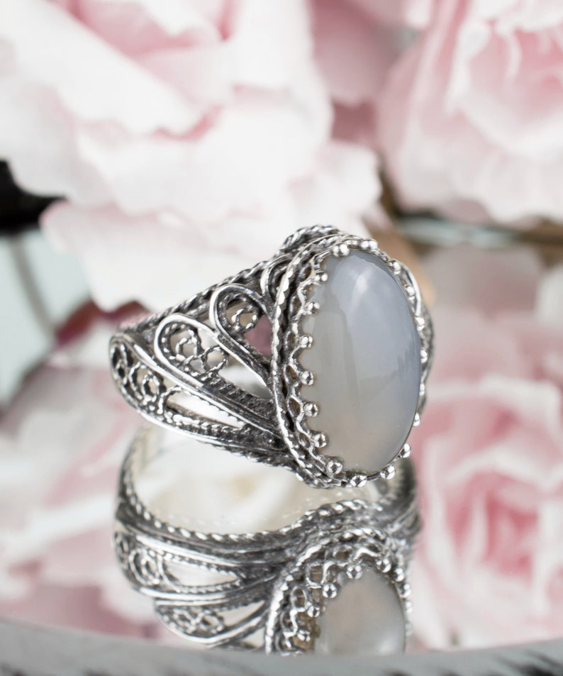 May include: A silver ring with an oval, gray gemstone. The ring has an ornate, filigree design around the band and the gemstone setting. The ring is reflected in a mirrored surface, with soft pink flowers in the background.