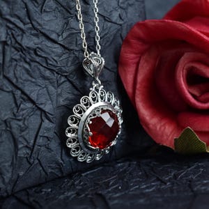 May include: A silver filigree pendant necklace with a red gemstone. The pendant is in the shape of a circle with a delicate design. The necklace is on a black background.
