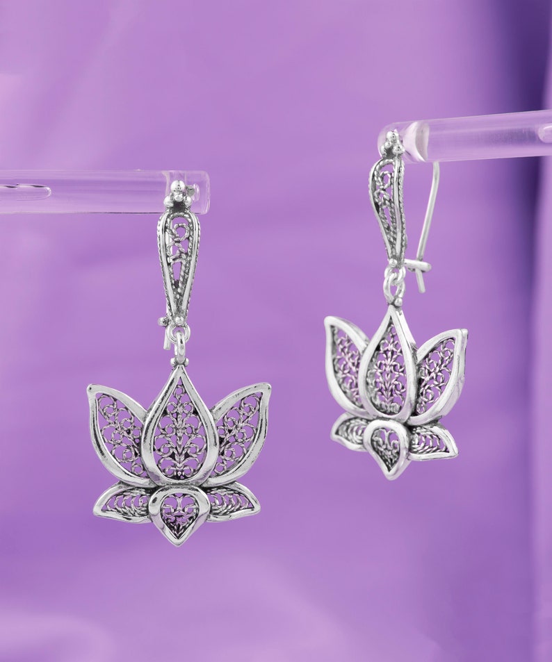 May include: A pair of silver filigree earrings with a lotus flower design. The earrings are hanging from a silver hook.
