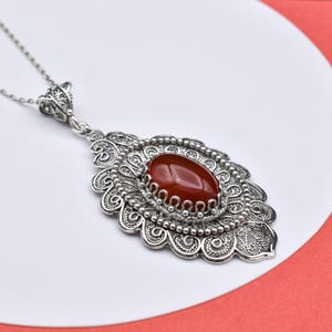 May include: A silver pendant necklace featuring an oval, reddish-brown gemstone set in an ornate, filigree design. The pendant is suspended from a delicate silver chain. The necklace is displayed on a white surface with a red background.