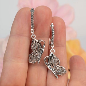 May include: A pair of silver filigree butterfly earrings with a delicate, intricate design. The earrings are hanging from a silver hoop.