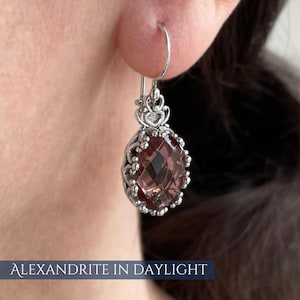 Op de afbeelding: Een zilveren oorbel met een facetgeslepen, ovaalvormige, roodbruine edelsteen. De edelsteen is gezet in een decoratief, opengewerkt frame met een hartvormig ontwerp. De tekst "ALEXANDRITE IN DAYLIGHT" staat onderaan.