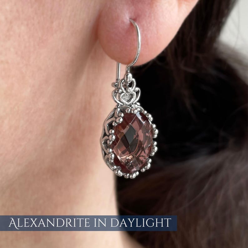 Op de afbeelding: Een zilveren oorbel met een facetgeslepen, ovaalvormige, roodbruine edelsteen. De edelsteen is gezet in een decoratief, opengewerkt frame met een hartvormig ontwerp. De tekst "ALEXANDRITE IN DAYLIGHT" staat onderaan.