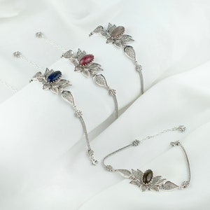 May include: Three silver bracelets with delicate filigree designs and oval-shaped gemstones. The gemstones are a light gray, a deep pink, and a dark blue.
