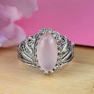 May include: A silver ring with an oval, light pink gemstone. The ring features an ornate filigree design around the gemstone and band. The gemstone is set in a beaded bezel. The ring is displayed on a wooden surface with a pink background.