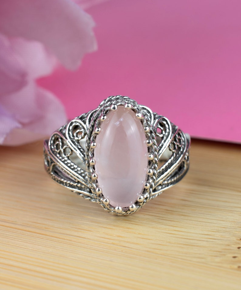 May include: A silver ring with an oval, light pink gemstone. The ring features an ornate filigree design around the gemstone and band. The gemstone is set in a beaded bezel. The ring is displayed on a wooden surface with a pink background.