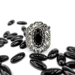 May include: A silver ring with a black oval gemstone set in a filigree design. The ring is surrounded by black oval-shaped beads.