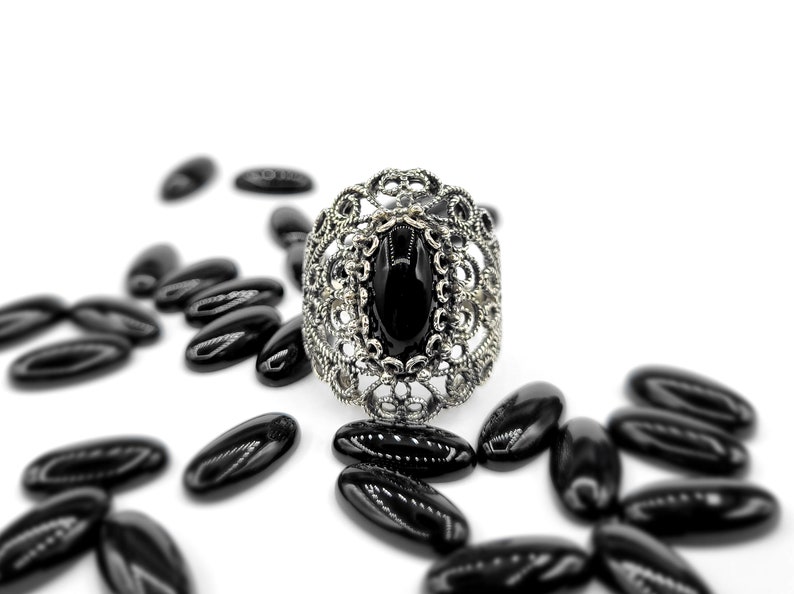 May include: A silver ring with a black oval gemstone set in a filigree design. The ring is surrounded by black oval-shaped beads.