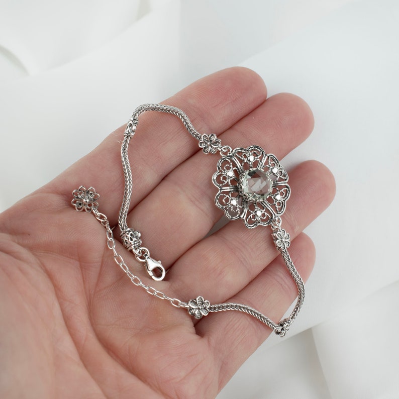 May include: A silver bracelet with a delicate chain and a large, ornate flower pendant featuring a clear gemstone in the center.