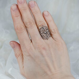 May include: A silver filigree ring with a delicate floral design. The ring is worn on the index finger of a hand.