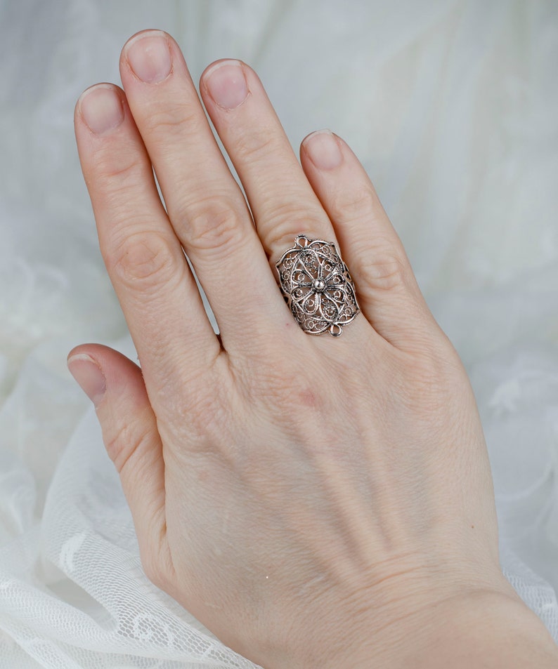 May include: A silver filigree ring with a delicate floral design. The ring is worn on the index finger of a hand.